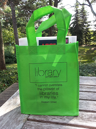 Lime green small tote bag with wording "library barrington area I cannot overstate the power of libraries in my life, quote from Elizabeth Gilbert" Bag is sitting on a wooden table, trees in the backgrouind
